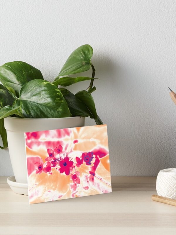 An art board print of a digitally altered photo of a Hypericum plant, in shades of reds, pinks, and peach. The art board print is leaning against the pot of a plant.